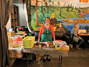 Joan Youngberg of the Columbia Arts Guild stands behind a table with wooden houses and paint for an art project. 