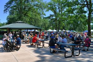 Citizens Day in the Park 2023 crowd at tables eating and standing next to covered pavilion in park