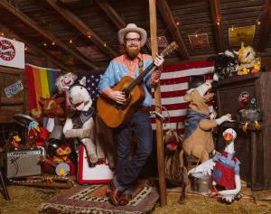 Man wearing western clothing in barn, smiling with guitar