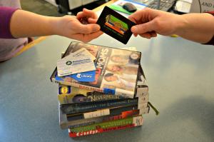 Stack of books with several Passport Program library cards on top and two hands passing a St. Helens library card. 