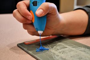 A closeup of a person's hand using a 3D pen to draw with a blue line. 